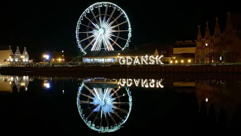 Nocny widok na podświetlony napis Gdańsk oraz diabelski młyn nad rzeką Motławą.