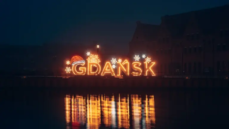 Podświetlony napis Gdańsk nad wodą w nocy przy historycznych zabudowaniach miasta.